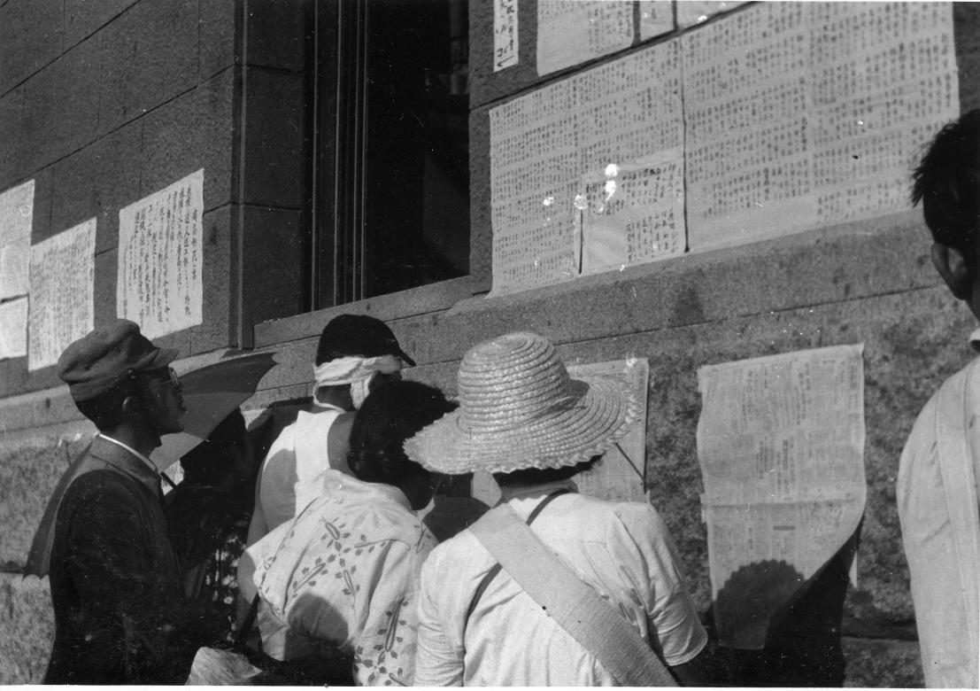 Citizens of Hiroshima and Nagasaki reading postings of a Japanese newspaper on a brick wall outside 