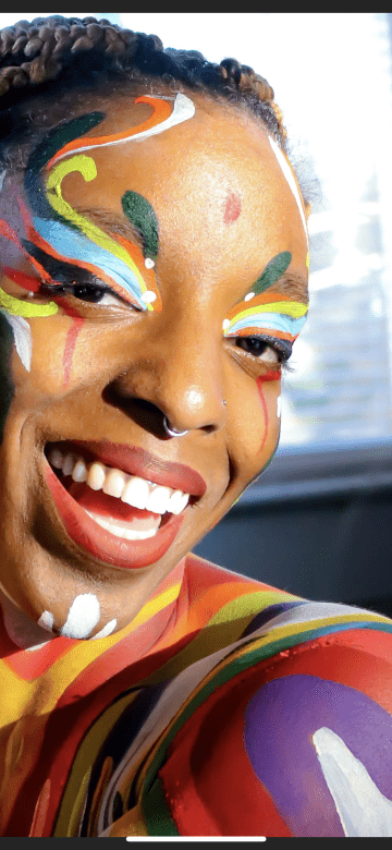 A photo of a black woman with different colors of face paint on her forehead, chin, cheeks, shoulders and collarbone. Some of the colors are purple, red, yellow, white, blue, green and orange.