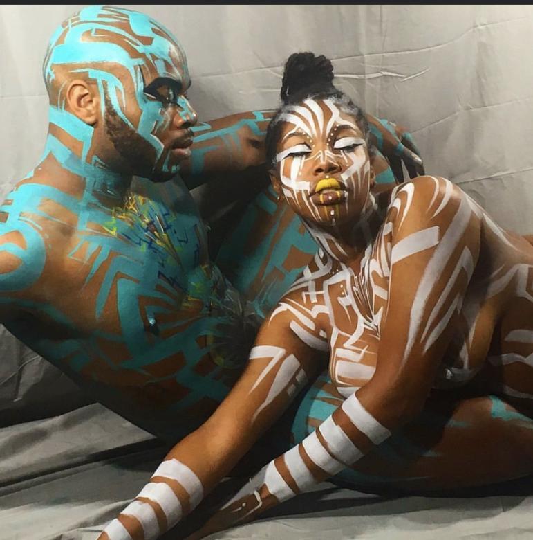 Image of a black man (left) sitting with eyes closed facing the right, his body and face is painted with blue and yellow paint. A black woman (right) is laying across the man with her hands in the foreground stretched out. Her eyes are closed and her face and body are painted with white stripes and dots featuring yellow paint on her lips.  