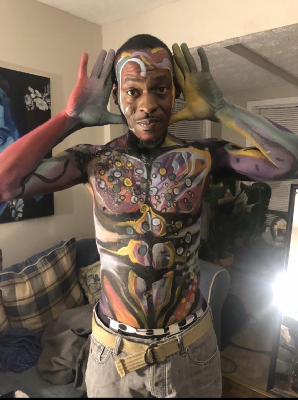 A photo of a black man with different colors of face paint on his body. Some of the colors include white, yellow, blue, purple, orange and grey.