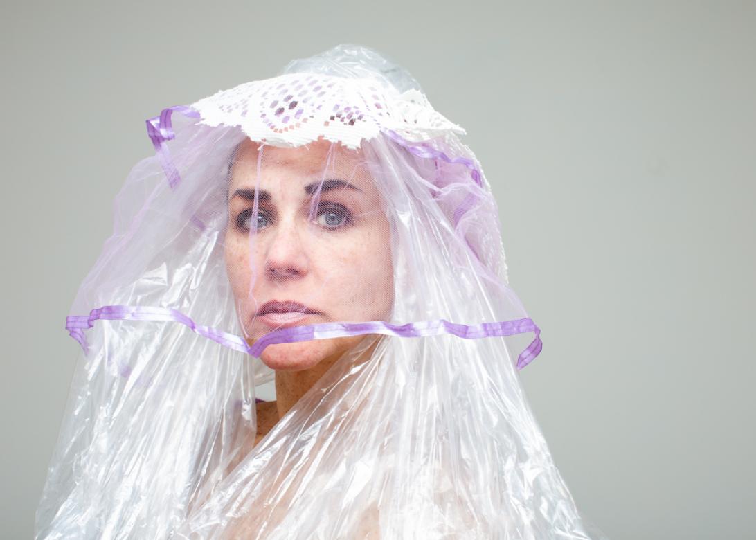 A photo of Eileen Powers faced to the left but looking to the viewer. She is wearing a long and layered headpiece with white lace on the top, cascading into light purple mesh with a purple ribbon trim over her face, and clear plastic coming down to each side of her chest.