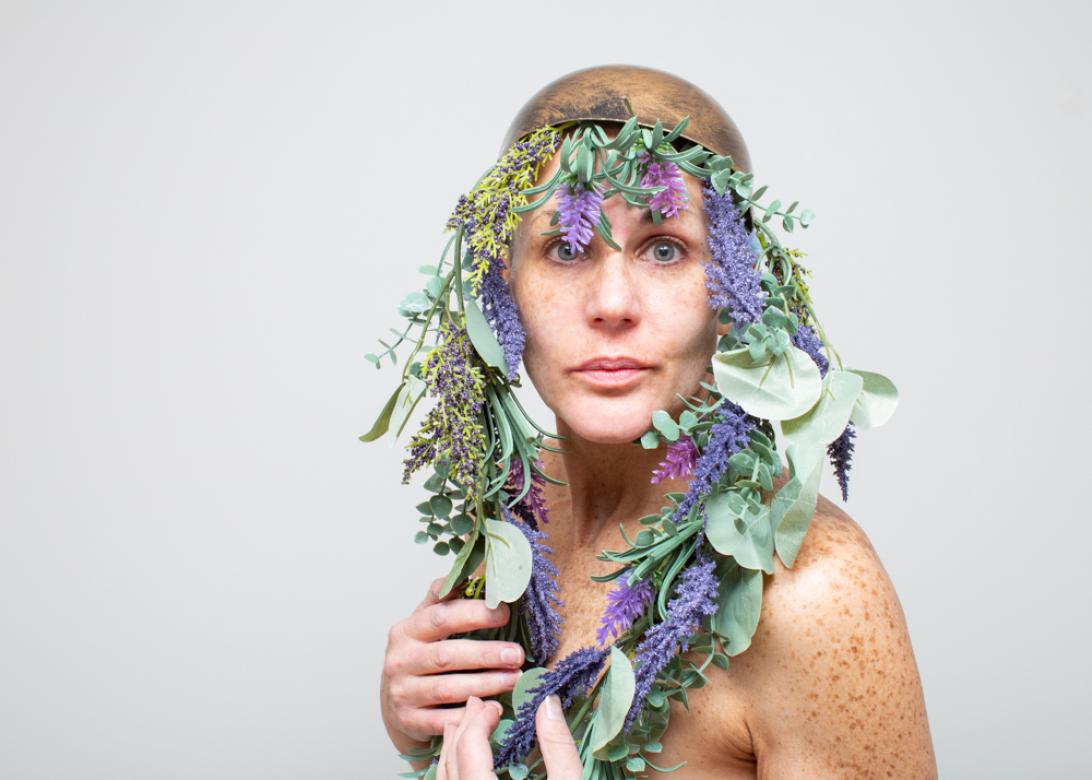 Photo of Eileen Powers wearing a brushed bronze cap with a garland of fake Lavender, Eucalyptus, Yellow and Purple Flowers, as well as other green across the front of the cap and hanging down each side of Eileen's face. Eileen clutches the garlands against her chest and looks at the viewer. 