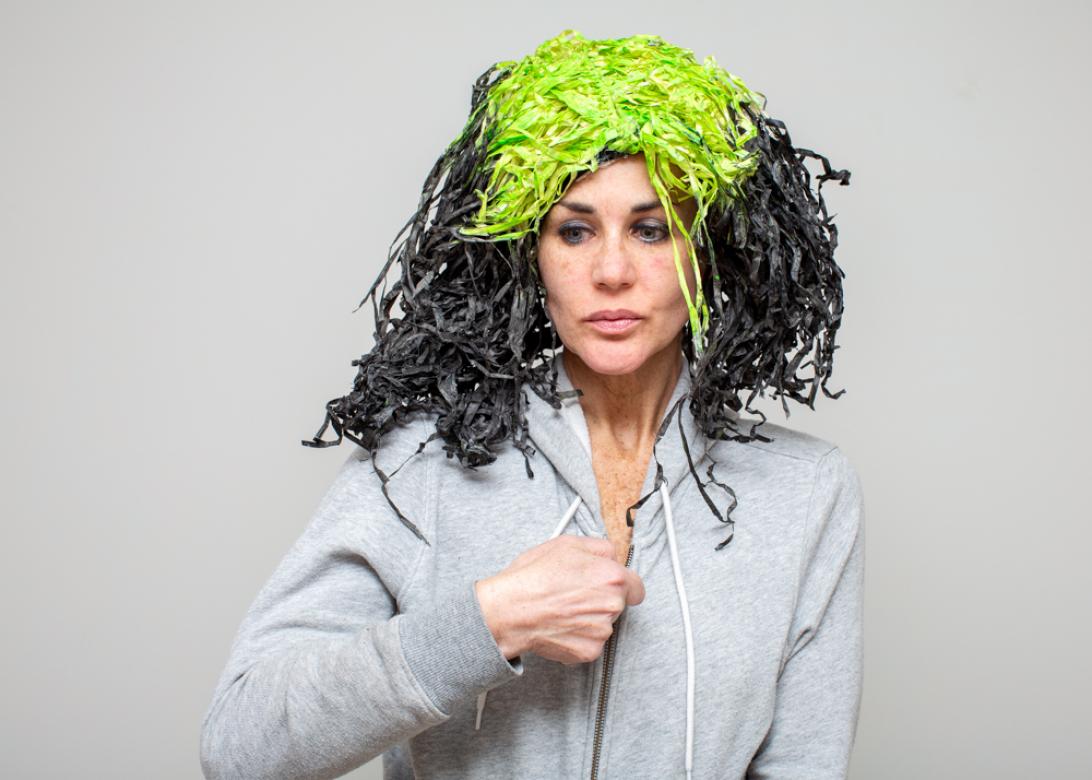 Photograph of Eileen Powers wearing a wig of black straw material to look like hair with lime green roots. She is looking down to the left and zipping up a grey sweatshirt. 