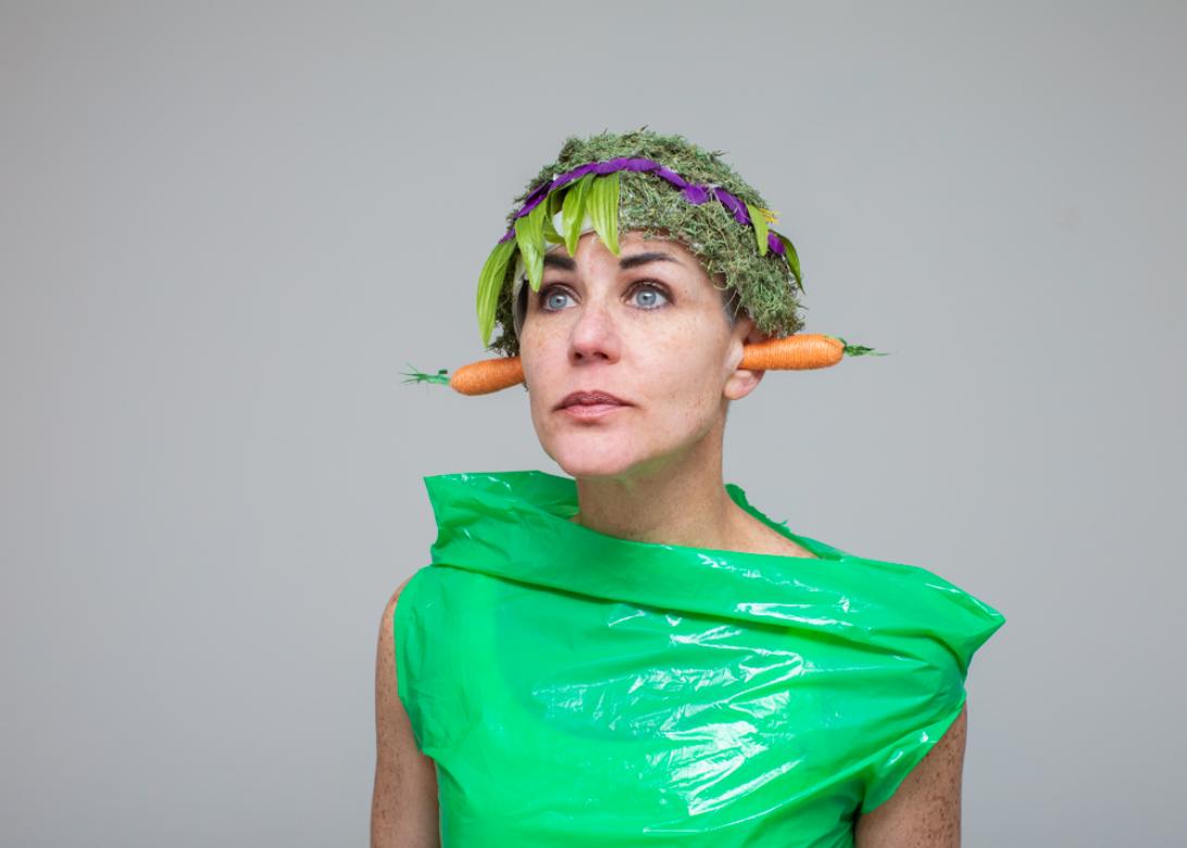 Photo of Eileen Powers facing towards and looking left wearing a bright green plastic bag as a top. She has a short headpiece on made of moss with a line of purple petals across with a line of light green leafs hanging down a bit. In each of Eileen's ear is a small straw carrot.