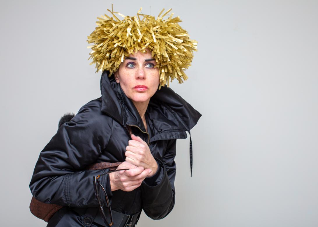 Image of Eileen Powers looking and facing to the right holding a bag and sunglasses with both hands. She is dressed in a navy blue jacket with a popped collar. She has a short wig sticking straight up made of golden yellow tissue strings.