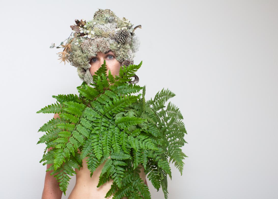 Photo of Eileen Powers staring at the camera with her hat tilted to the left. She is covering her chest with large ferns leafs that also cover the bottom half of her face and nose. Her eyes are fixed on the viewer with a tilted eyebrow. Her hair piece is a short but voluminous piece made from light colored mosses, yellow thistles, dried brown leaves, and pinecones. 