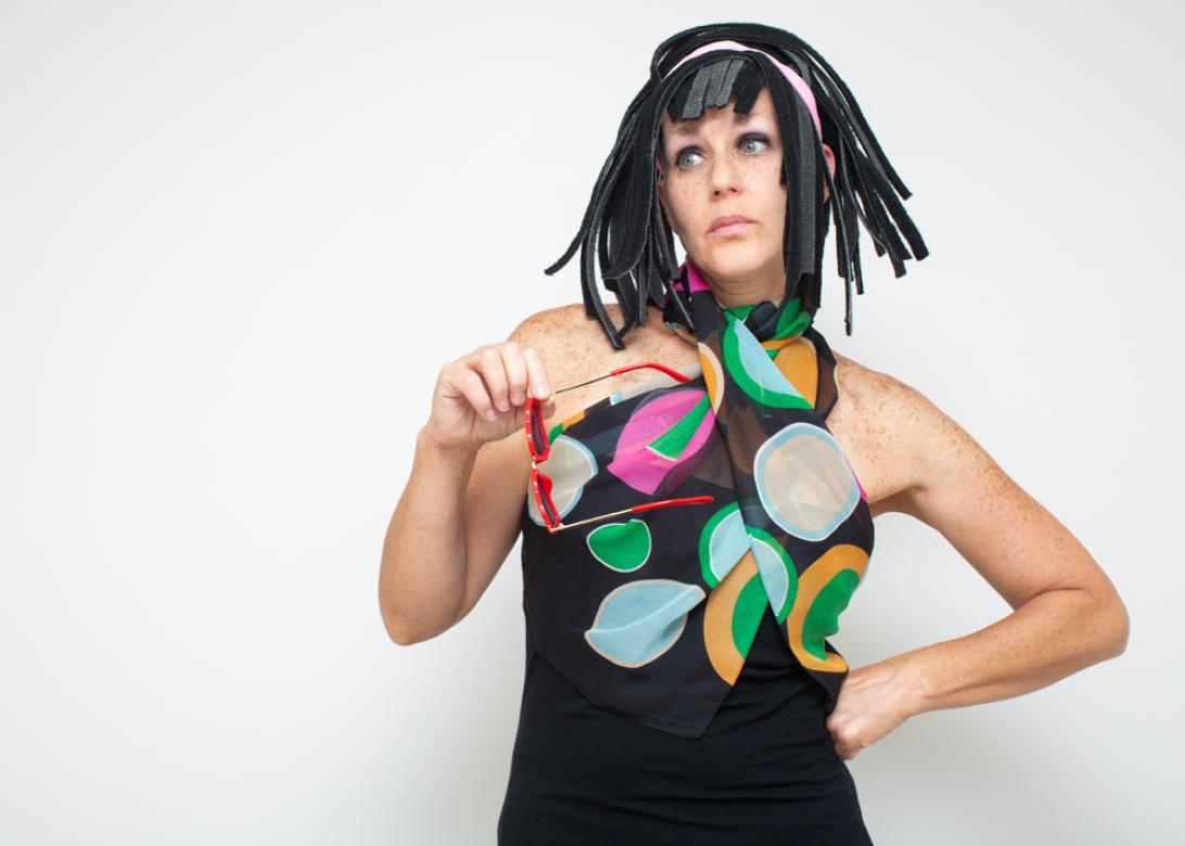 Photo of Eileen powers looking right with her left hand on her hip. In her right hand is a red pair of sunglasses. She is in a black dress with a black scarf around her neck with brightly colored large polka dots. Her hair is a flippy black wig with bangs that is shoulder length and made of black car mats. The wig features a thin pink headband. 