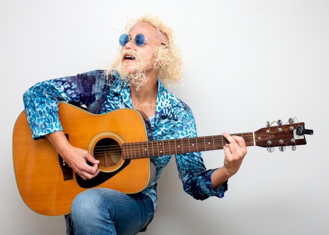 A Photo of Eileen Powers wearing a blue batik long sleeve button up, with jeans and round blue 60s style sunglasses. She is sitting facing the left holding and playing an acoustic guitar while singing.  She has medium length blonde wig made from straw packing material with an accompanying mustache and beard.
