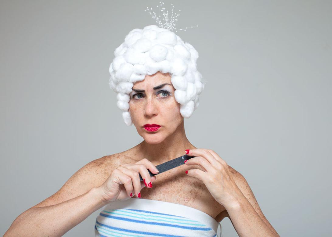 Photo of Eileen Powers looking to the right with her eyes raise and eyebrow raised. She is holding her left hand up with red nail polish on her fingers while filing them with the other hand. She has on a blue and white striped tube top, her bright red lipstick matches her nails. Her wig is a short wig made from cotton balls in the style of a powdered wig. At the top of the wig  little white beaded strings are bursting out straight up. 