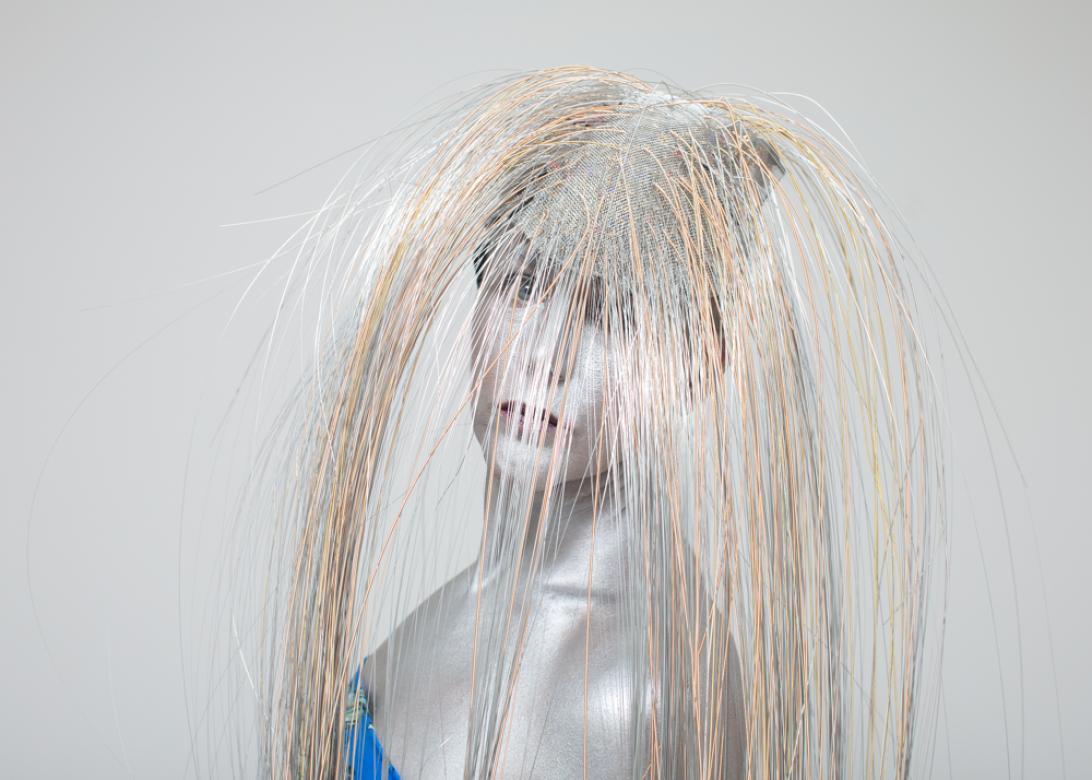 In this photo of Eileen Powers her body and face is painted a metallic silver, while she wears a blue tube top. Her body is angled left while she turning her head looking straight at the viewer. She has a long voluminous wig made from instrument strings that are silver and bronze. The hairs come out of a silver mesh base.