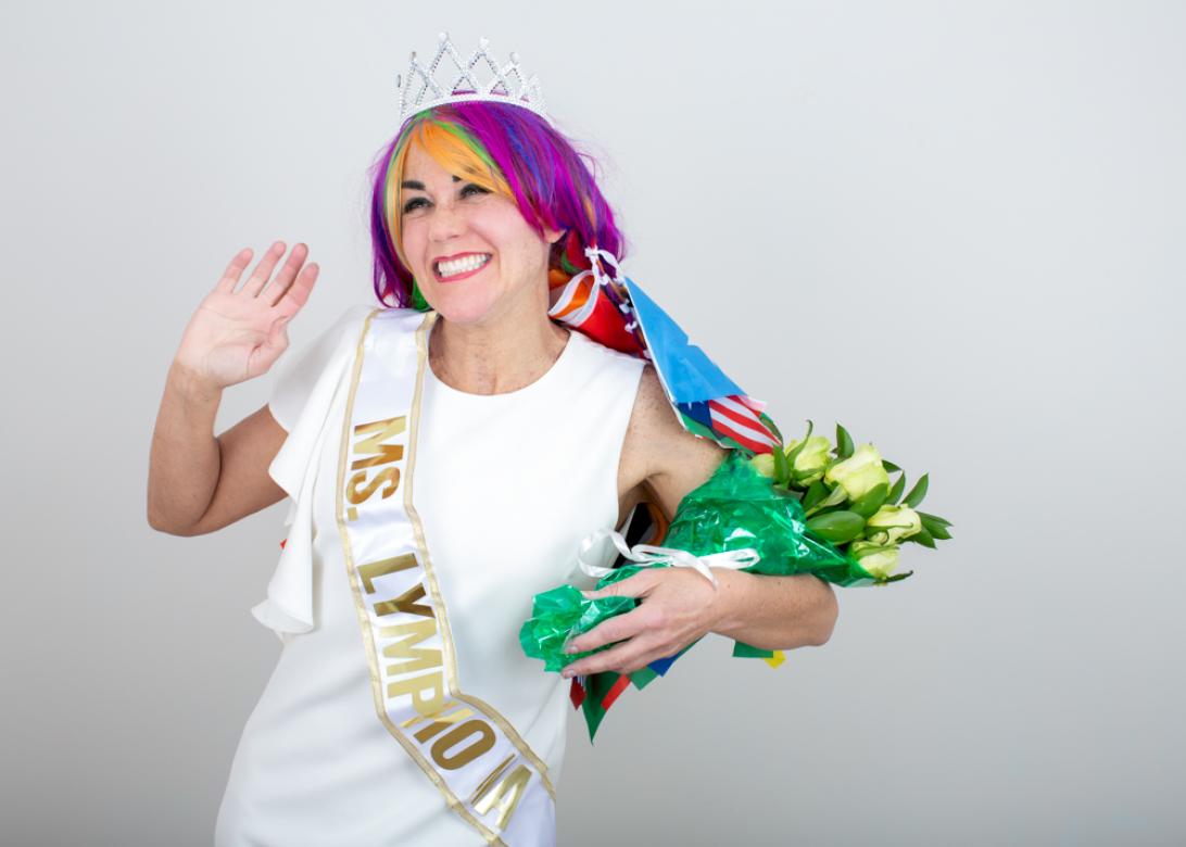 In this photo of Eileen Powers she is facing left waving happily smiling and clutching a bouquet in the style of a newly crowned beauty queen. She is wearing a white dress with a white and gold sash that read "miss lymphoma". Her hair is quite long comprised of rainbow hair extensions at the tops with many small flags from different countries at the bottom. She has a silver crown on top. 