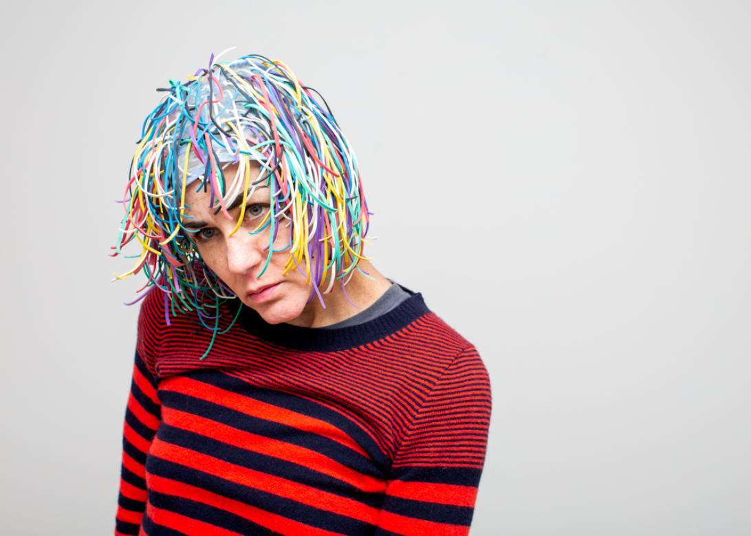 A photo of Eileen Powers towards the left of the frame leaning in with her head tilted as she stares into the distance in a moody way. She is wearing a navy and red striped sweater with a medium length fringey wig with long bangs made from different color rubber bands. 