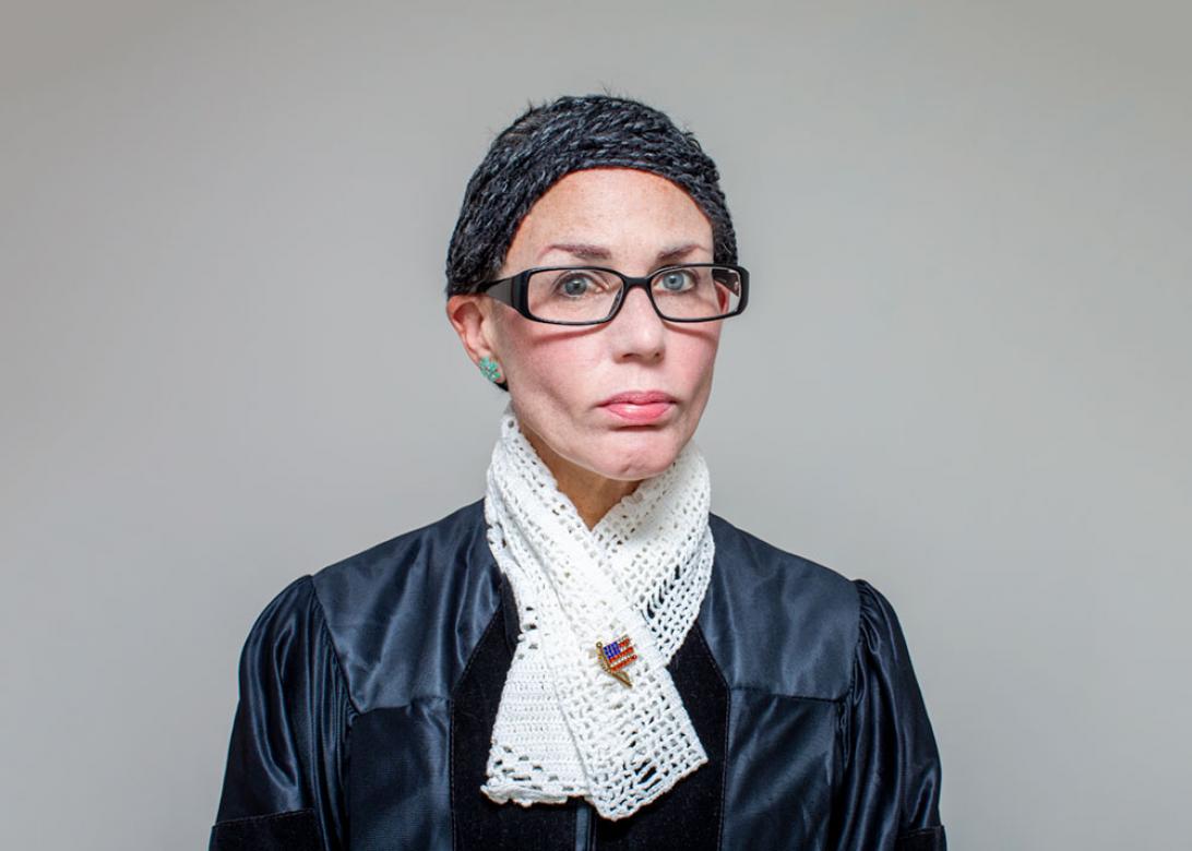 Photo of Eileen Powers facing and looking directly at the viewer, wearing a judges robe with a short white crocheted scarf with an American flag pin. She is wearing short black grey hair made from yarn slicked back. She also has on glasses and green flower shaped earrings with pink lipstick. 
