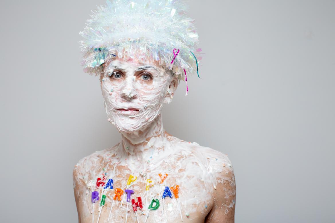 In this photo of Eileen Powers she facing and looking right at the viewer with her hands at her side towards her back. She is covered in frosting on her chest, neck, and face. On her chest candles in rainbow colors spell out "happy birthday". She is wearing a short but puffy wig made of iridescent white holiday tinsel.  