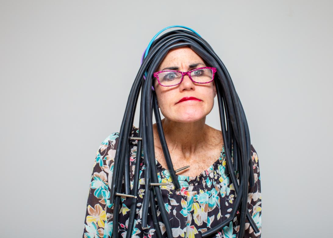 A photo of Eileen Powers facing the viewer looking towards the right. She is wearing a multicolored floral print long sleeve dress, she also has on hot pink leopard print glasses with hot pink lipstick to match. She has on a long wig made of bike tubes complete with inflation nozzles. She has a thin blue and purple headband with the hair. She is looking stumped biting her lips.