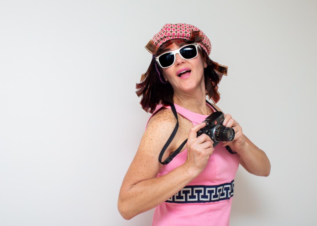 A photo of Eileen Powers moving towards the right holding a film camera up pressing the shutter, the camera is on a black lanyard around her neck.. She is smiling widely wearing pink lipstick and white sunglasses. She has on a 60s style light pink dress with a printed belt at the midsection. She has a flippy styles should length wig made from brown film strips with bangs. She is wearing a deep pink, blue, and cream color printed beret. 