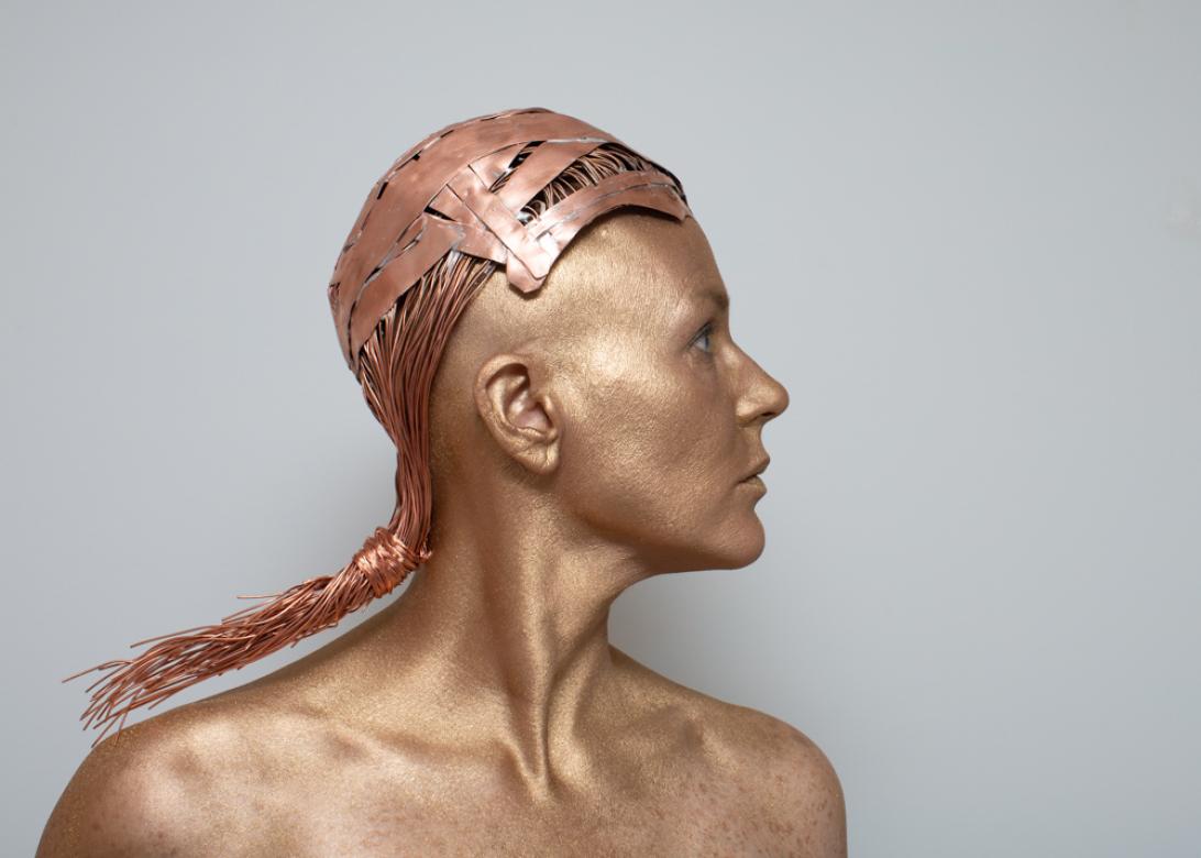 In the side profile photo of Eileen Powers her body is facing the viewer with her head entirely turned to the right. She is covered in a metallic copper body paint. Her wig is made from larger copper parts at the front transitioning into long skinny copper wires that form a slicked back pony tail secured at the base of EIleen's neck.