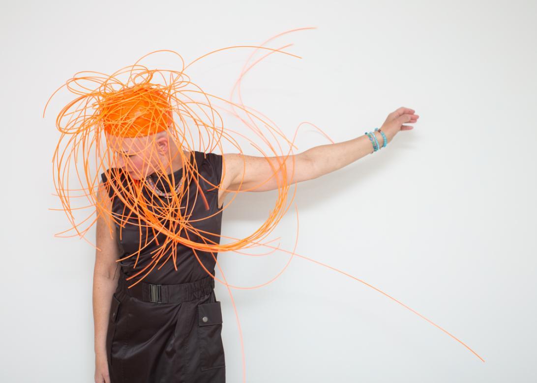 In this knee up portrait of Eileen Powers she is positioned to the left of the frame looking down holding her left arm up with an open hand. She is wearing shiny black cargo style pants with a matching belt and black tank top. She has on a multicolored thin beaded necklace, with three turquoise beaded bracelets on her raised hands. Her headpiece is a bright orange pillbox style cap fit snug with long orange plastic strings jutting out from every angle, some forming loops and swooping in front of her face.