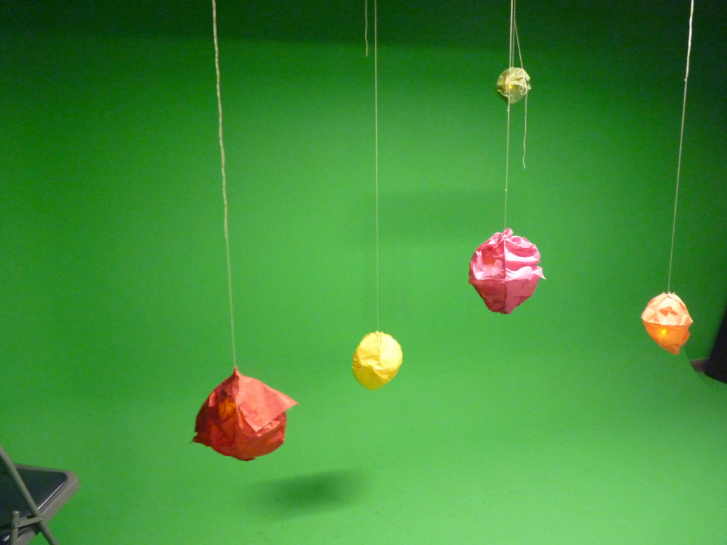 Colored balls, which appear to be made from tissue paper, hang from the ceiling in a green-screened room.