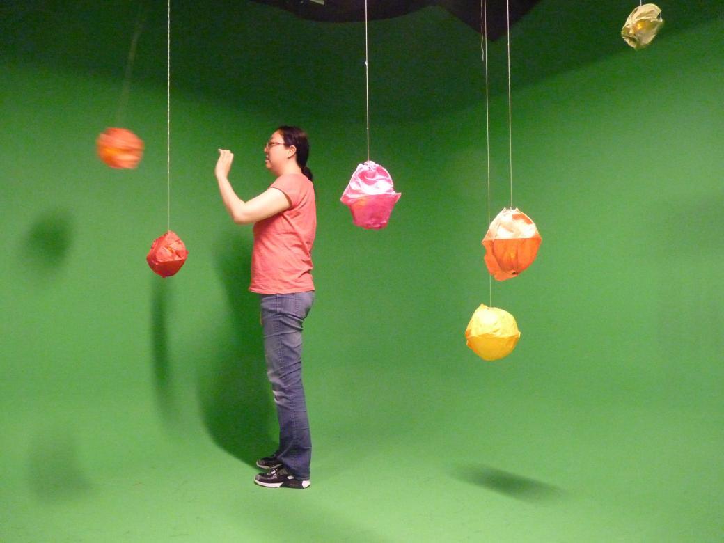 A student stands in the green-screened room with the hanging colored balls. The student's hand is outstretched as she had just pushed one of the balls.