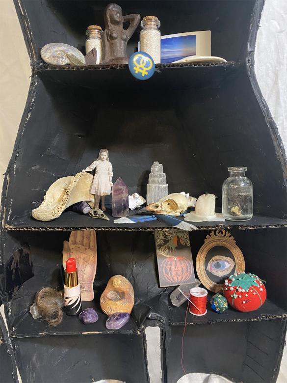 close-up of left half of mannequin, bottom half, containing trinkets such as feathers, a candle, and lipstick