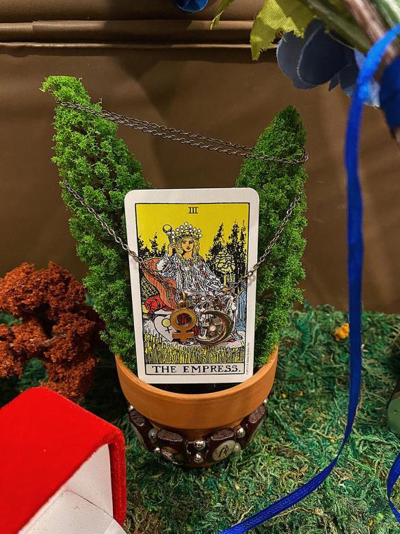 Close-up of tarot card that says "The Empress" in a pot with mini trees and a necklace