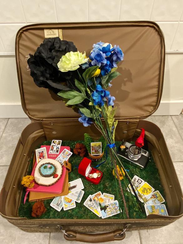 Open suitcase that contains flowers, notebooks, a watch, a camera, tarot cards, and other trinkets