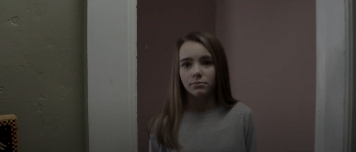 Young girl standing in a hallway looks sadly off-screen.