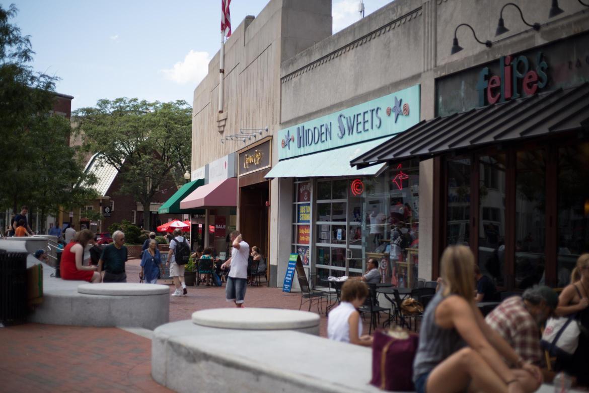 Row of restaurants and shops in Harvard Square. Some people are walking by, some are sitting.