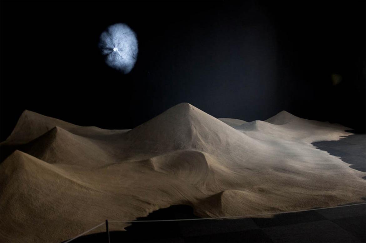 A large area of sand sculpted into desert dunes in the corner of a dark room. A bright, circular, swirly object is projected on the wall above the dunes.