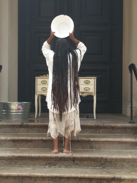 A black person with long dreadlocks faces away towards a large black door, a white desk, and a metal bucket. They are lifting up a white bowl and are pouring water onto their head.