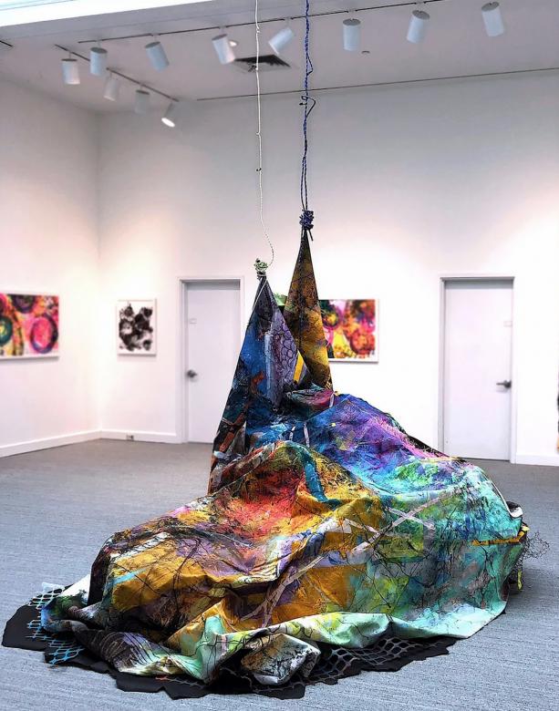 A large, mountainous rainbow-colored sheet sits in the middle of an art gallery. Two of its corners are attached to wires that come down from the ceiling.