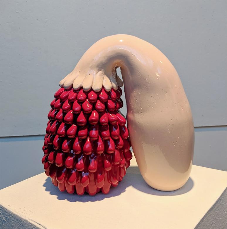 A ceramic sculpture. A beige squash-like object bends down, sprouting red blob-like petals that reach the ground.