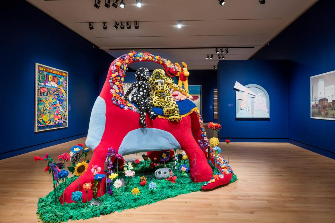 A large colorful and playful soft sculpture of two figures covered in polka dots sitting on a smiling red dinosaur.