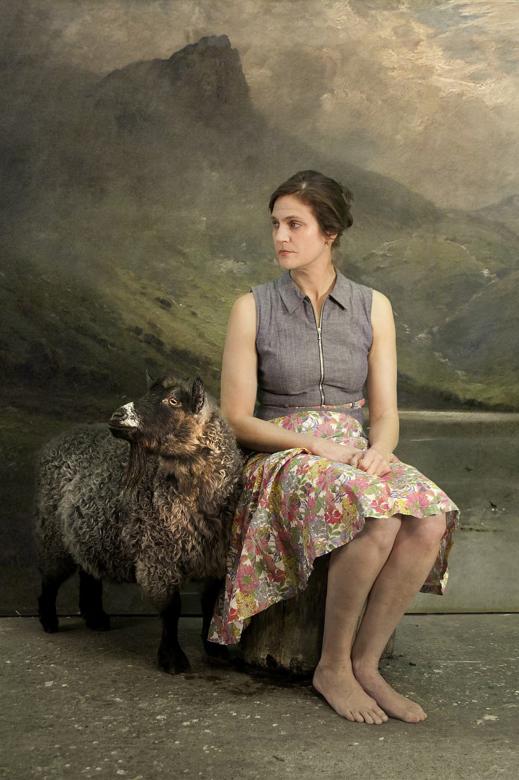 A woman sits on a stump in front of a painted mountainous landscape. A brown sheep stands next to her. Both are looking to the left.