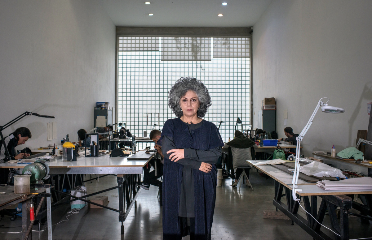 Doris Salcedo stares defiantly at the camera while crossing her arms. She stands in the center of an art studio where other people are bent over desks, working.