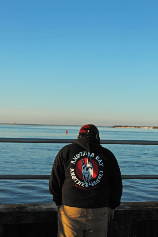Person wearing a red cap and a black hoodie facing a body of water. The back of the hoodie says "Another day, another regret."