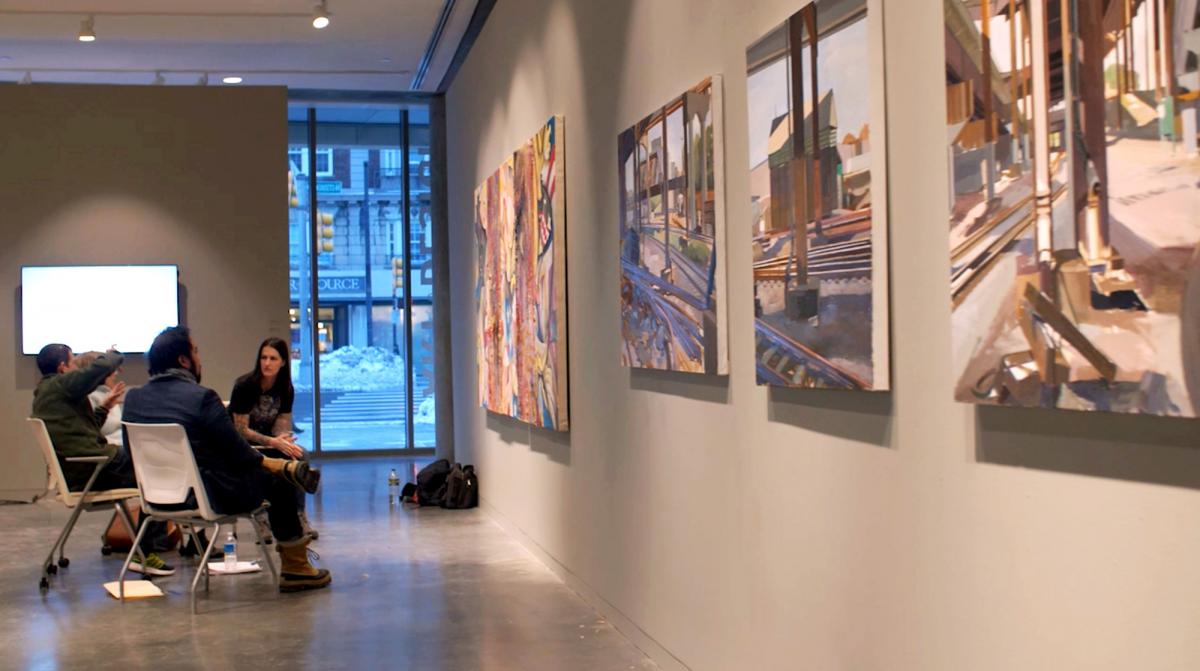 Student and faculty sitting in chairs having a discussion in front of the student's work that hangs in a gallery.
