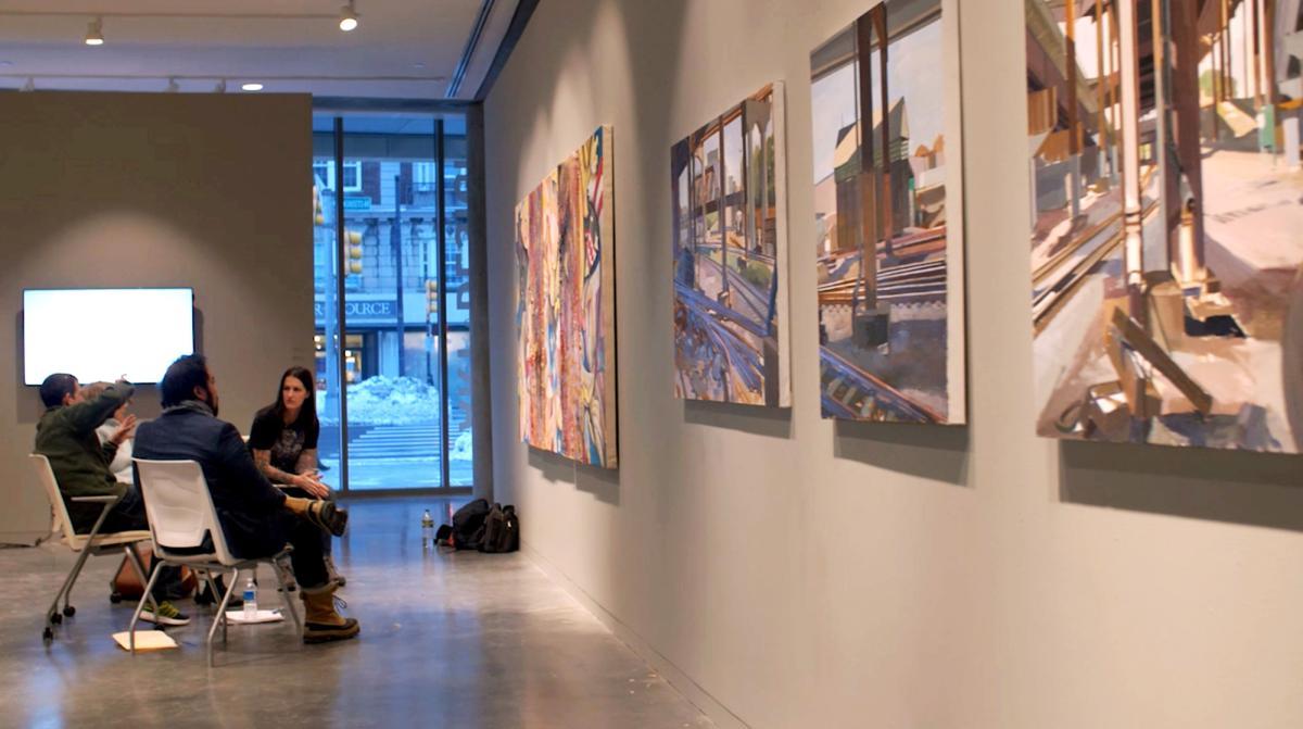 Student and faculty sitting in chairs having a discussion in front of the student's work that hangs in a gallery.