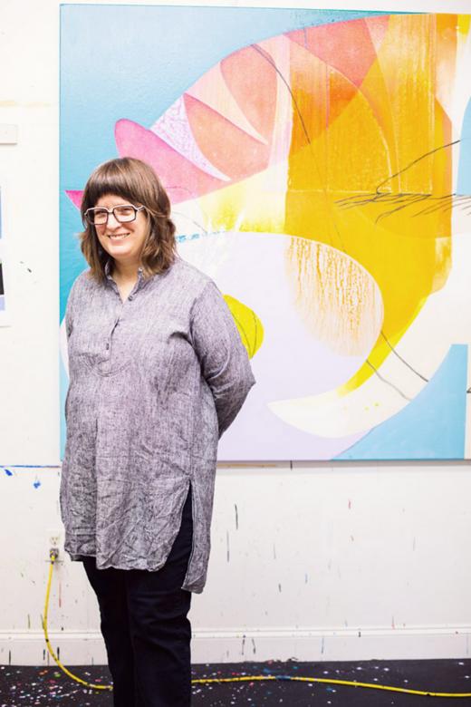 Carrie Moyer stands and smiles with her hands behind her back. She is in front of an abstract blue, pink, orange, yellow, and white painting.