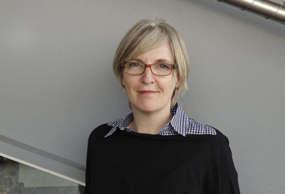 Helen Molesworth stands in front of a gray wall, wearing glasses and a black collared shirt.