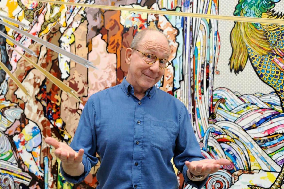 Jerry Saltz smiles and shrugs in front of a colorful, textured, wall. The wall appears to depict sun rays, rock formations, and waves.