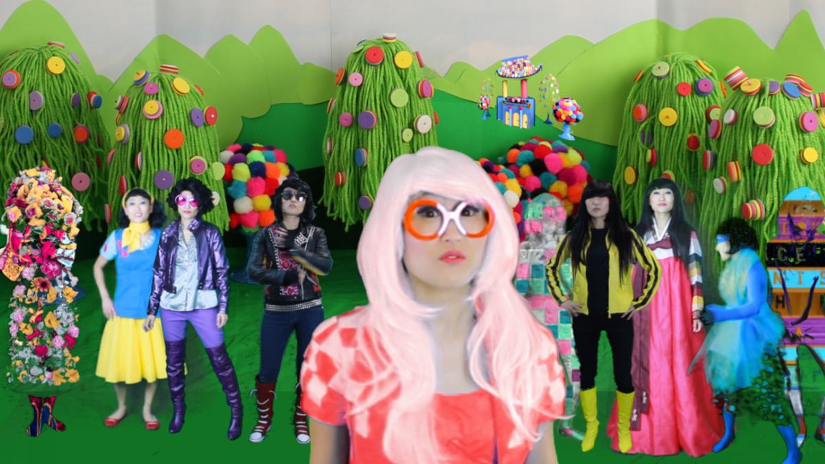 JooYoung Choi wearing a pink wig and round orange glasses stands in a fictional green world surrounded by herself in different costumes.