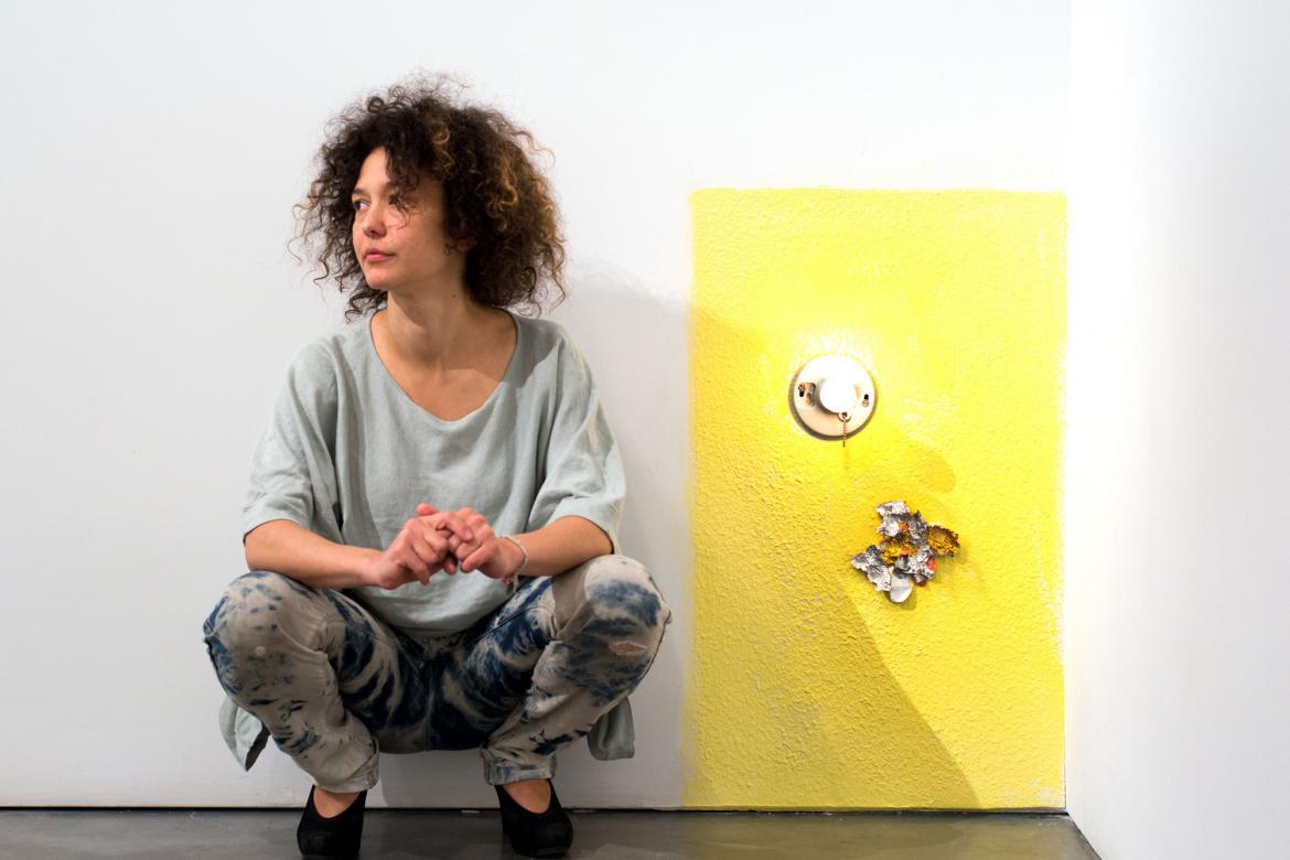 Mika Rottenberg squats in front of a white wall. Next to her, a small rectangular portion of the wall is painted yellow, with two metal objects stuck to it.