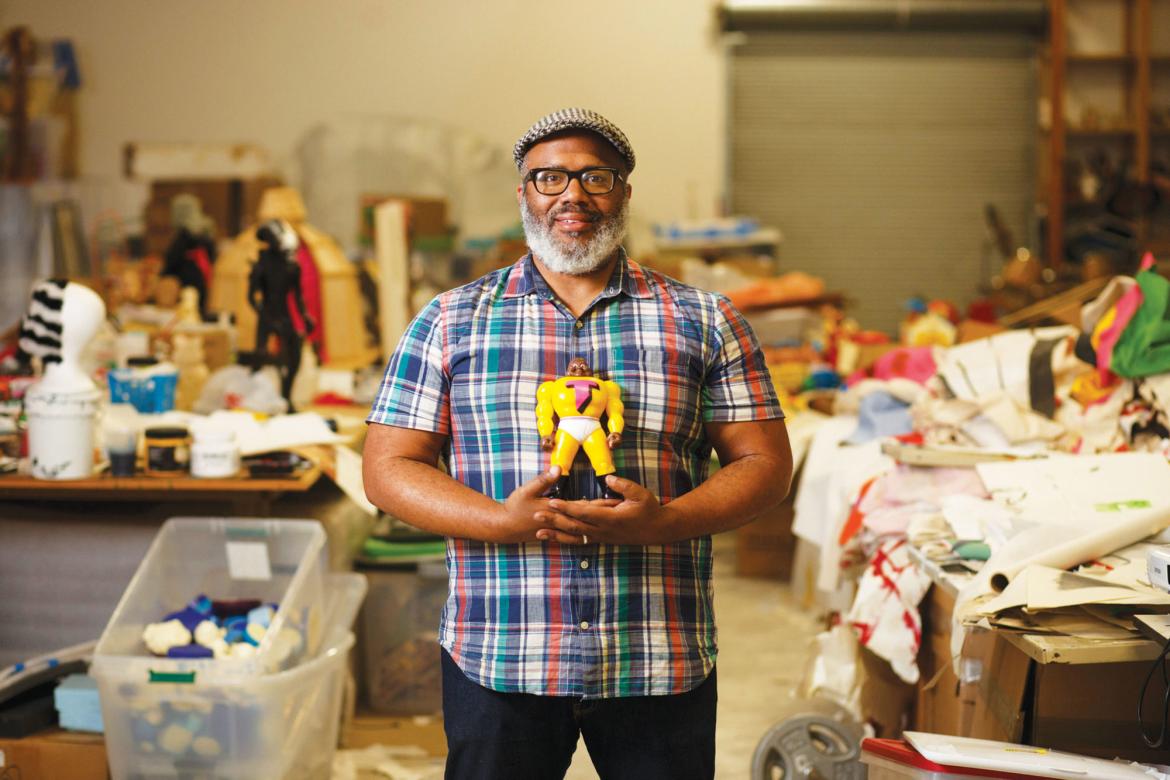 Trenton Doyle Hancock stands in an art studio messy with paper, wood, figurines, and other art materials. He smiles while holding a superhero figurine.