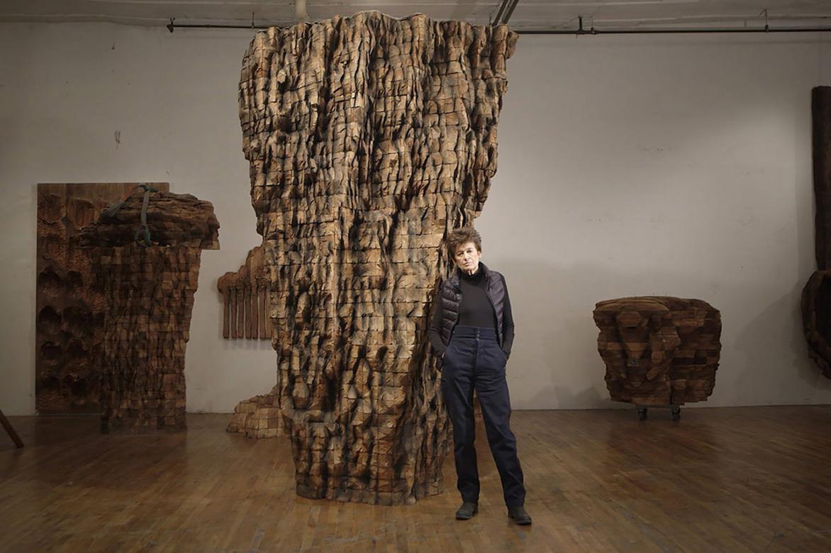 Ursula Von Rydingsvard stands in an art gallery surrounded by various large, abstract sculptures made from wood. 