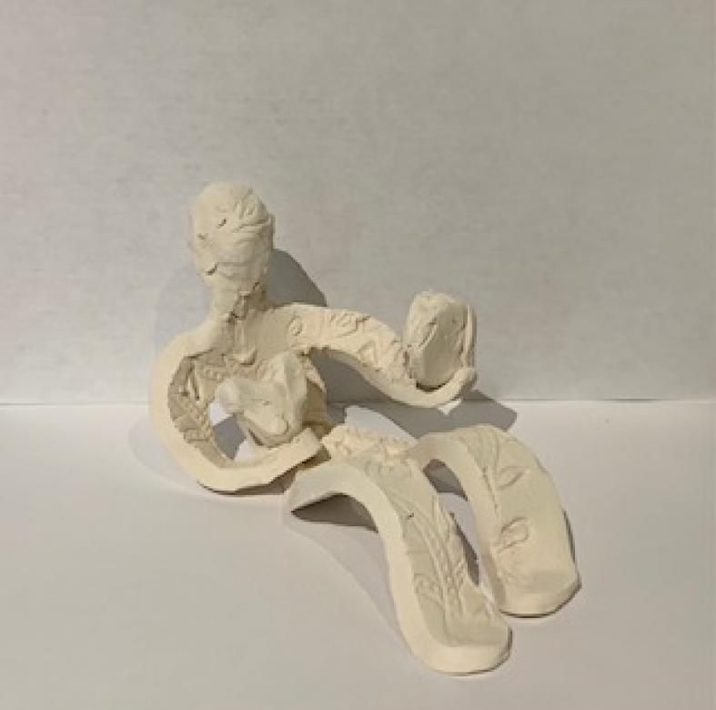 White clay sculpture of a figure sitting and holding a heart with one hand raised looking into a mirror. The figures body is embossed with patterns.