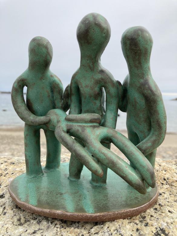 Small ceramic sculpture of three figural forms holding a fourth figure who is laying down with their hand on their heart. Each figure has their hand on the shoulder of the center figure. The piece is glazed in green and copper glaze. It is photographed on a rock at the beach.