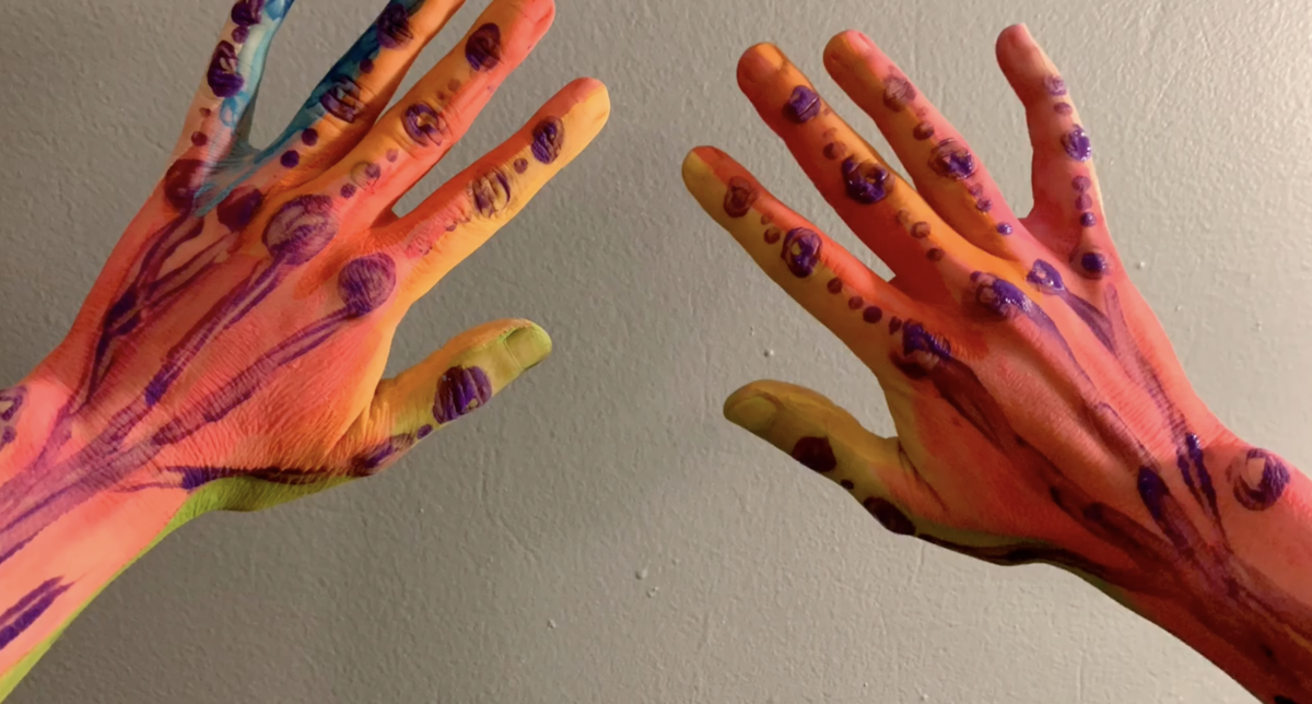 Artists two hands reaching out covered in pink, red, orange, and yellow paint. Purple paint lines go up the hands with a purple dot on each knuckle with smaller dots between. Some green and blue paint is between the fingers on the left hand. 
