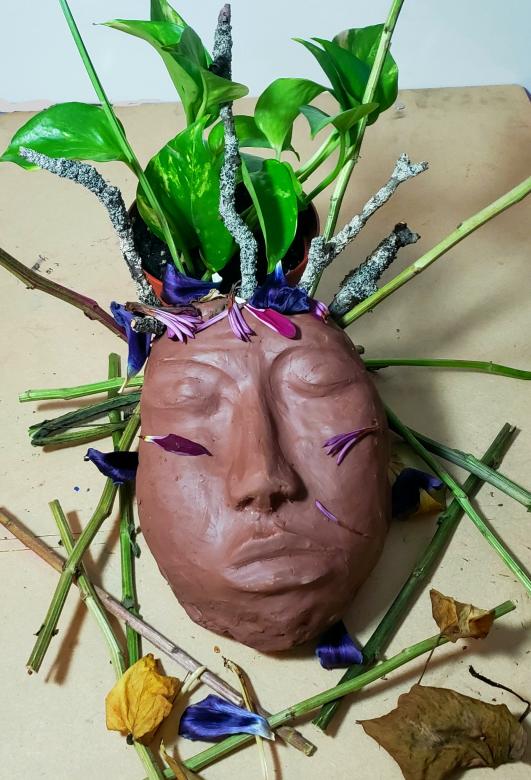 A terracotta colored clay sculpture of a face with green potho leaves sprouting from the top of the ahead along with sticks, flower petals, cut flower stems, and dried leaves.
